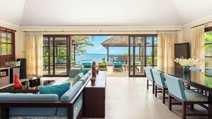 A spacious living room with a large sliding glass door leading to a balcony with a view of the ocean. There is a sofa set, a dining table with chairs, and a small kitchen area visible. - Luxury Beachfront Pool Villa - Finu@Samui (Koh Samui)
