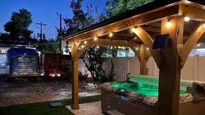 Un bain à remous sous une pergola en bois, illuminé de guirlandes lumineuses, entouré d’une zone recouverte de gravier et d’une pelouse.