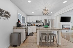 A modern kitchen with white cabinets, granite countertops, a central island with stools, a built-in oven, microwave, and a large flat-screen TV above the island.