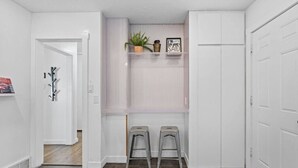 Un couloir avec un porte-manteau, deux tabourets, une plante et un panneau sur une étagère.