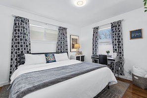 A bedroom with a bed, desk, chair, and a window with patterned curtains.