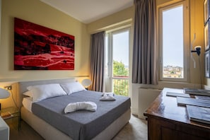 A hotel room with a bed, a desk, a window with curtains, and a painting on the wall.