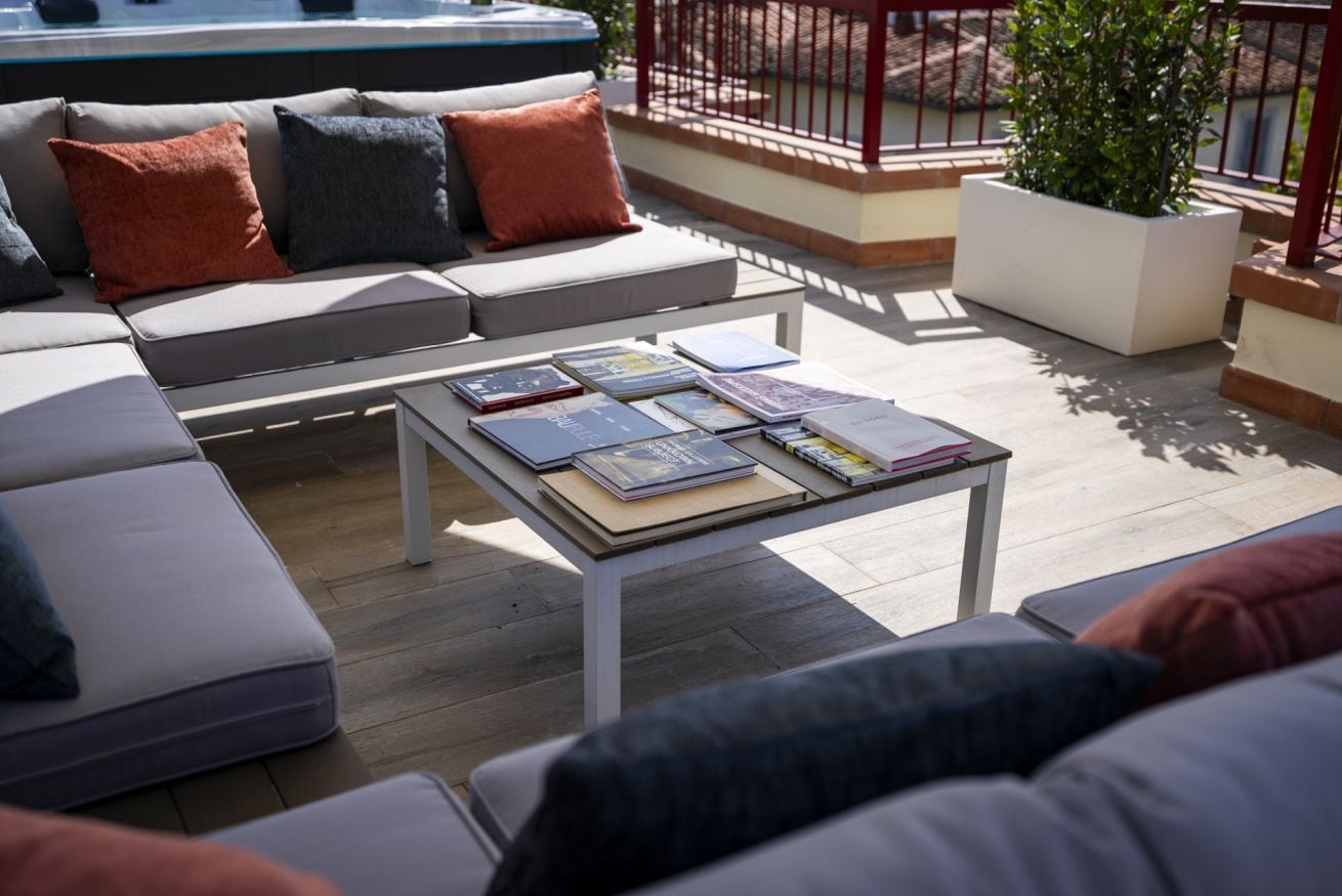 Un espace extérieur moderne aménagé avec un canapé, des coussins et une table sur laquelle sont posés des livres.