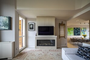A modern living room with a flat-screen TV, fireplace, large windows, and a dining area.