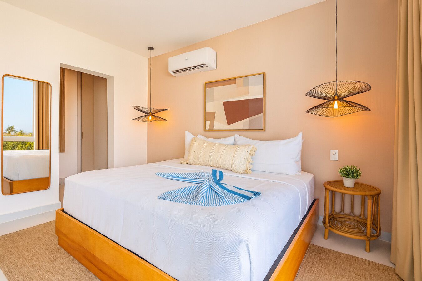 A hotel room with a large bed, a wall-mounted air conditioner, a framed abstract painting, a unique wall lamp, and a small round table with a plant.