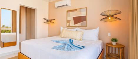A hotel room with a large bed, a wall-mounted air conditioner, a framed abstract painting, a unique wall lamp, and a small round table with a plant.