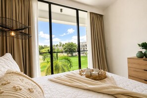 A hotel room with a large window, a bed with a white quilt, a wooden dresser, and a basket of toiletries. - Elegant 2BR Cocotal condo with golf views, pool access & prime Punta Cana location (Punta Cana)