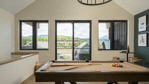 A pool table in a room with large windows offering a view of a residential area and mountains.