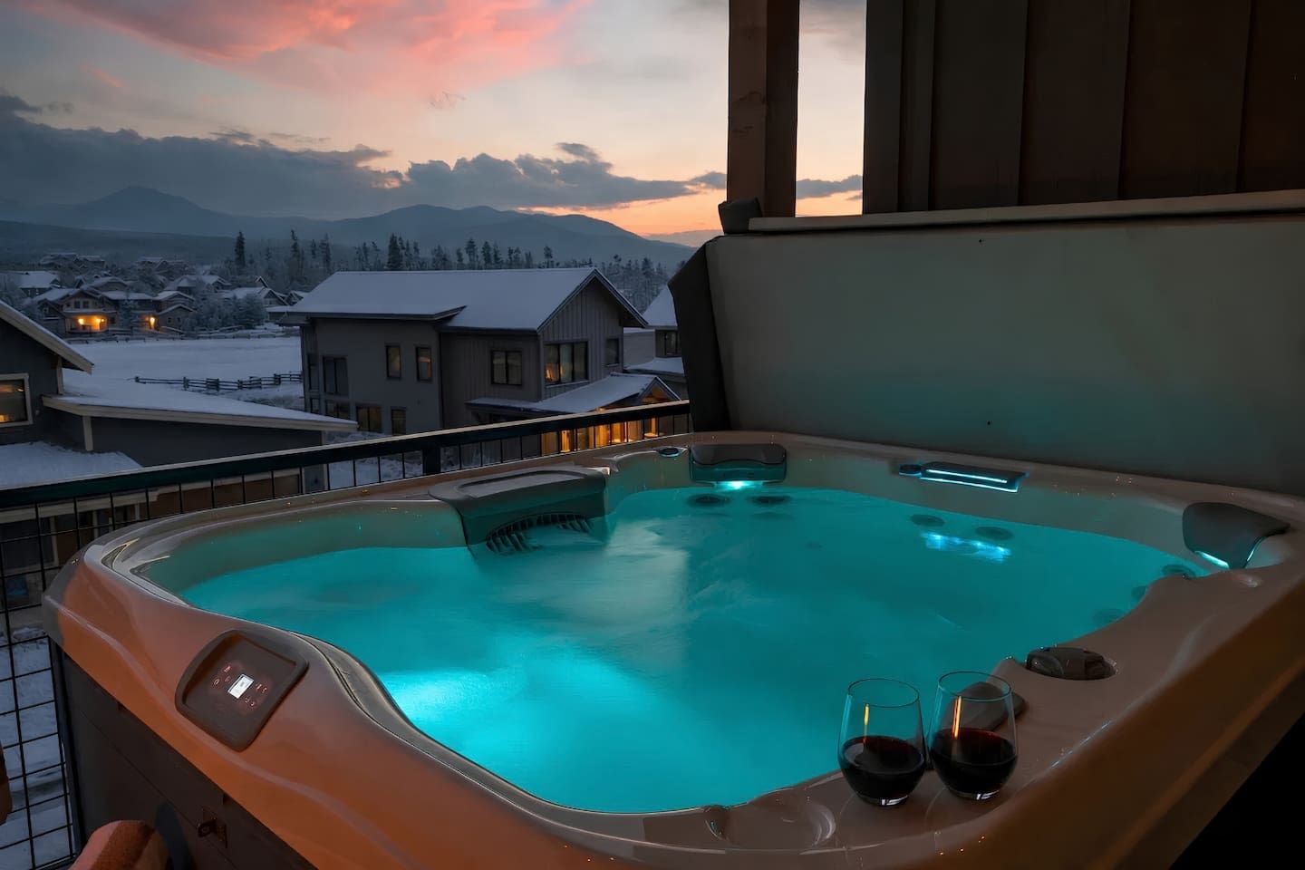 Un jacuzzi con dos copas de vino tinto en un balcón con vistas a un pueblo nevado.