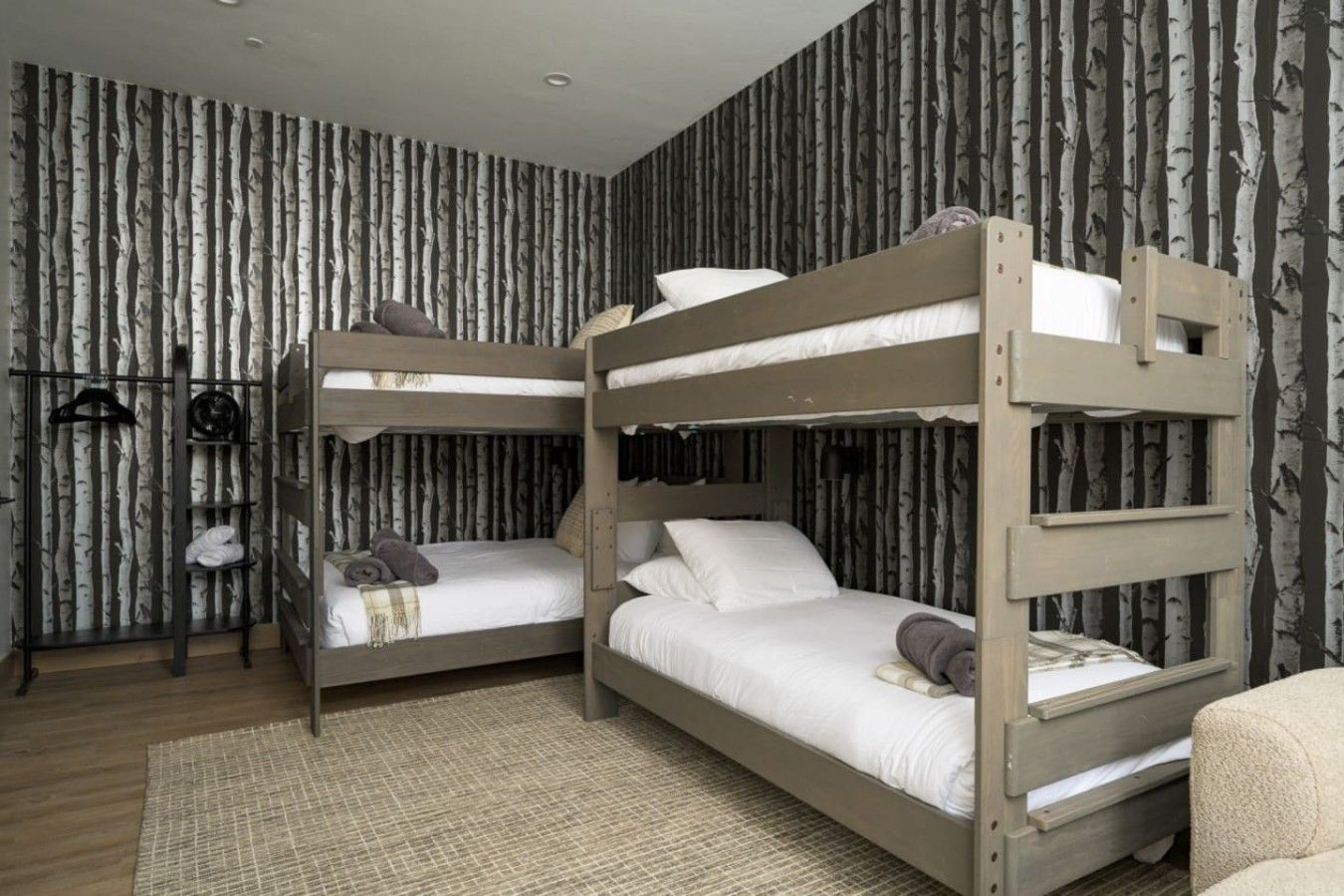 A room with two bunk beds, a wooden floor, a woven rug, and a wardrobe with hanging items.