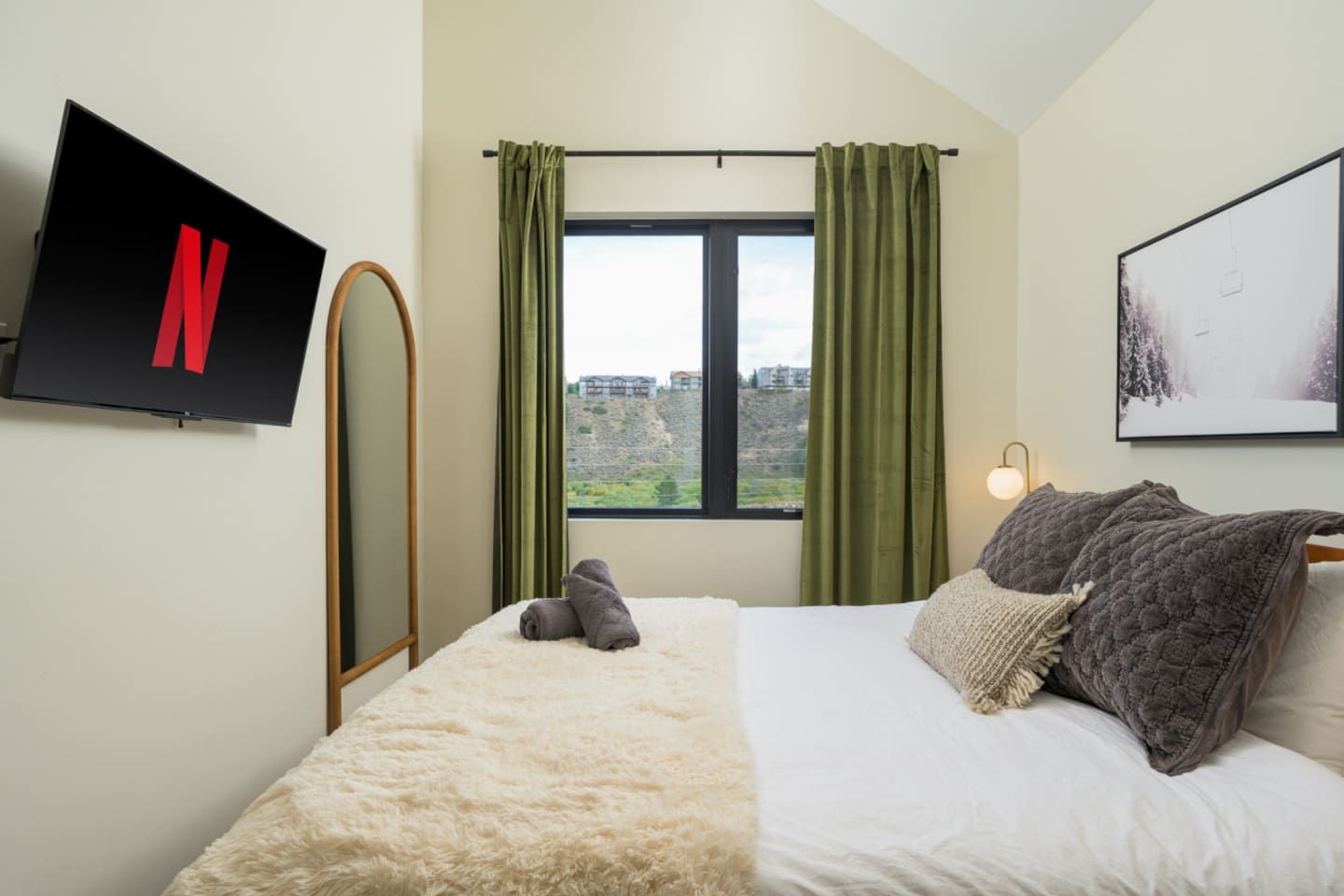 A hotel room with a bed, a TV mounted on the wall, a window with curtains, and a painting on the wall.
