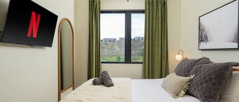 Habitación de hotel con cama, televisión montada en la pared, ventana con cortinas y un cuadro en la pared.