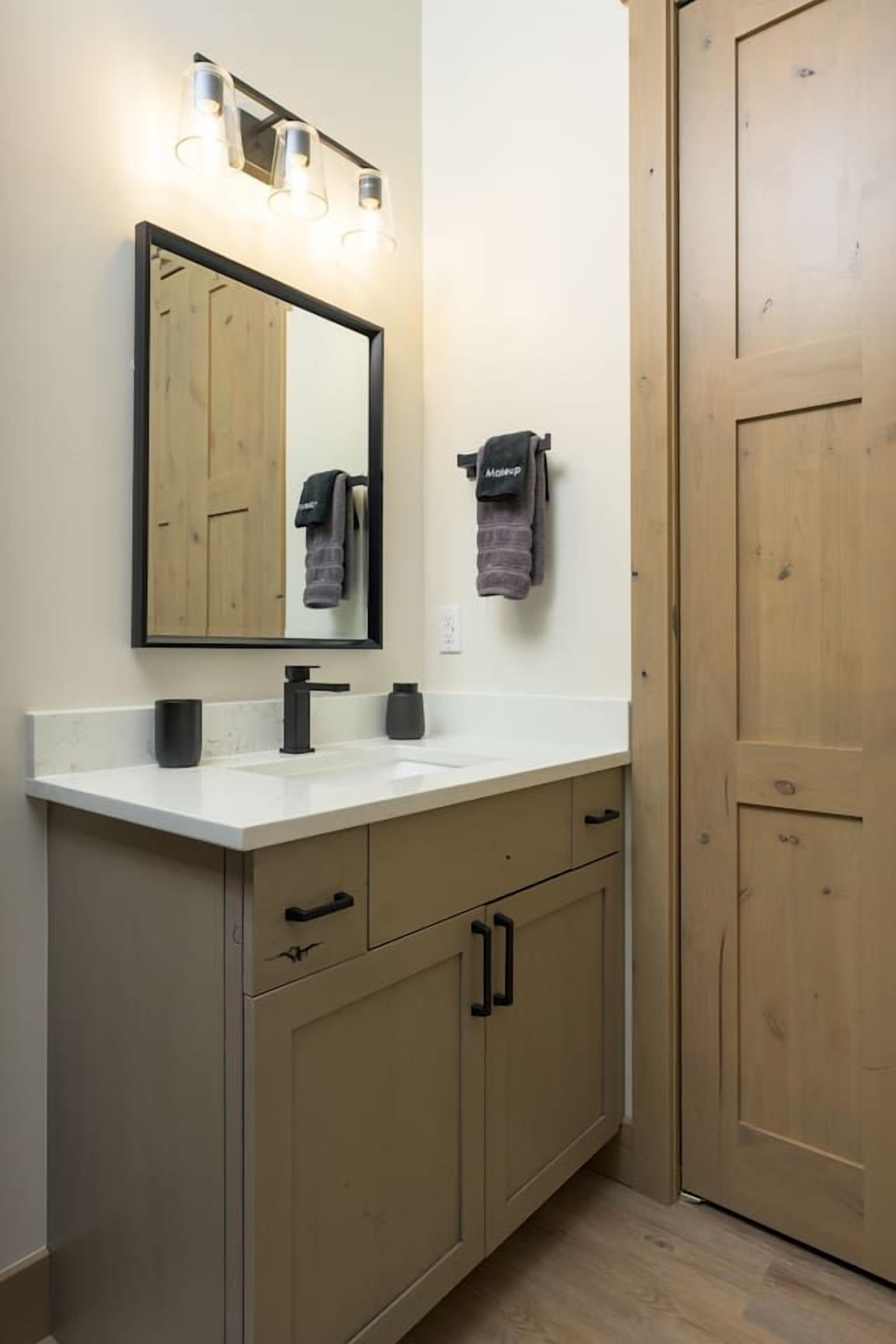 Een badkamer met een witte wasbak, een zwarte kraan en een spiegel met verlichting. Handdoeken hangen aan een rek en er is een houten deur.