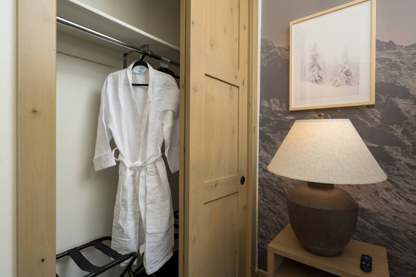Une porte en bois donne sur un placard où est suspendue une robe blanche. On aperçoit une lampe sur une table de chevet et un tableau encadré au mur.