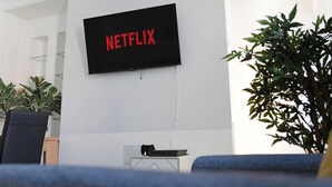Ein Wohnzimmer mit einem an der Wand montierten Fernseher, der 'NETFLIX' anzeigt, einer Spielkonsole und einer Pflanze.