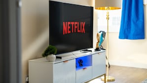 Un soggiorno moderno con una TV a schermo piatto che mostra il logo di Netflix, una lampada decorativa e una piccola pianta su un mobile bianco.