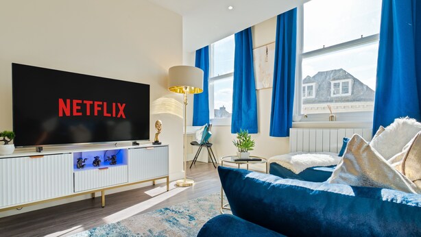 Un soggiorno moderno con una grande TV a schermo piatto che mostra "NETFLIX", un mobile bianco con oggetti decorativi, un divano blu e vista su una montagna attraverso ampie finestre con tende azzurre.