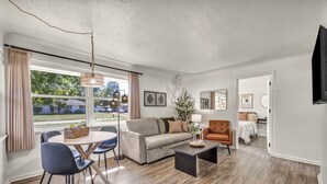 A modern living room with a sofa, armchairs, a coffee table, and a dining table. There is a large window with curtains, a fan, and a vase with flowers. The room has hardwood flooring and a flat-screen TV mounted on the wall.