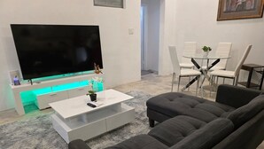 A modern living room with a large flat-screen TV, a white coffee table, a dark grey sofa, and a glass dining table with white chairs.