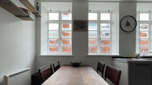 Comedor con mesa y sillas de madera, un radiador blanco y ventana con vistas a edificios.