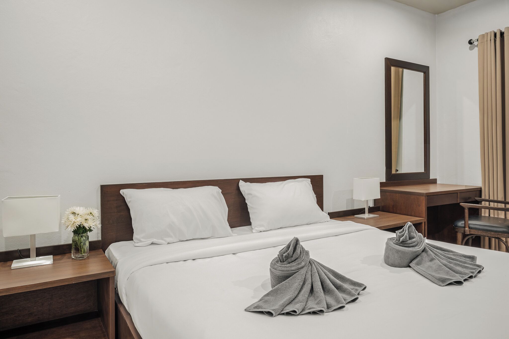 Una camera d'albergo con un letto rifatto, un comodino con lampada e fiori, una scrivania con sedia e uno specchio grande appeso al muro.