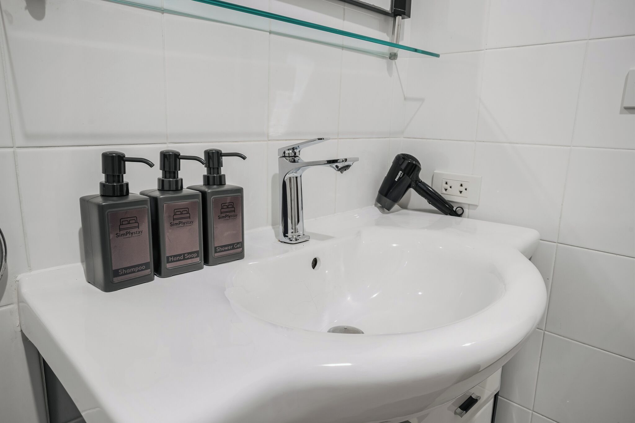 A bathroom with a white sink, a handheld showerhead, and three bottles of Simplesay shampoo and hand soap.