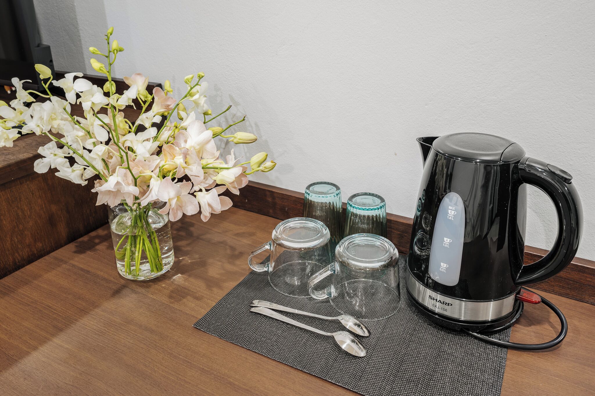 A wooden table with a black electric kettle, two glass mugs, a spoon, a fork, and a vase with white flowers.