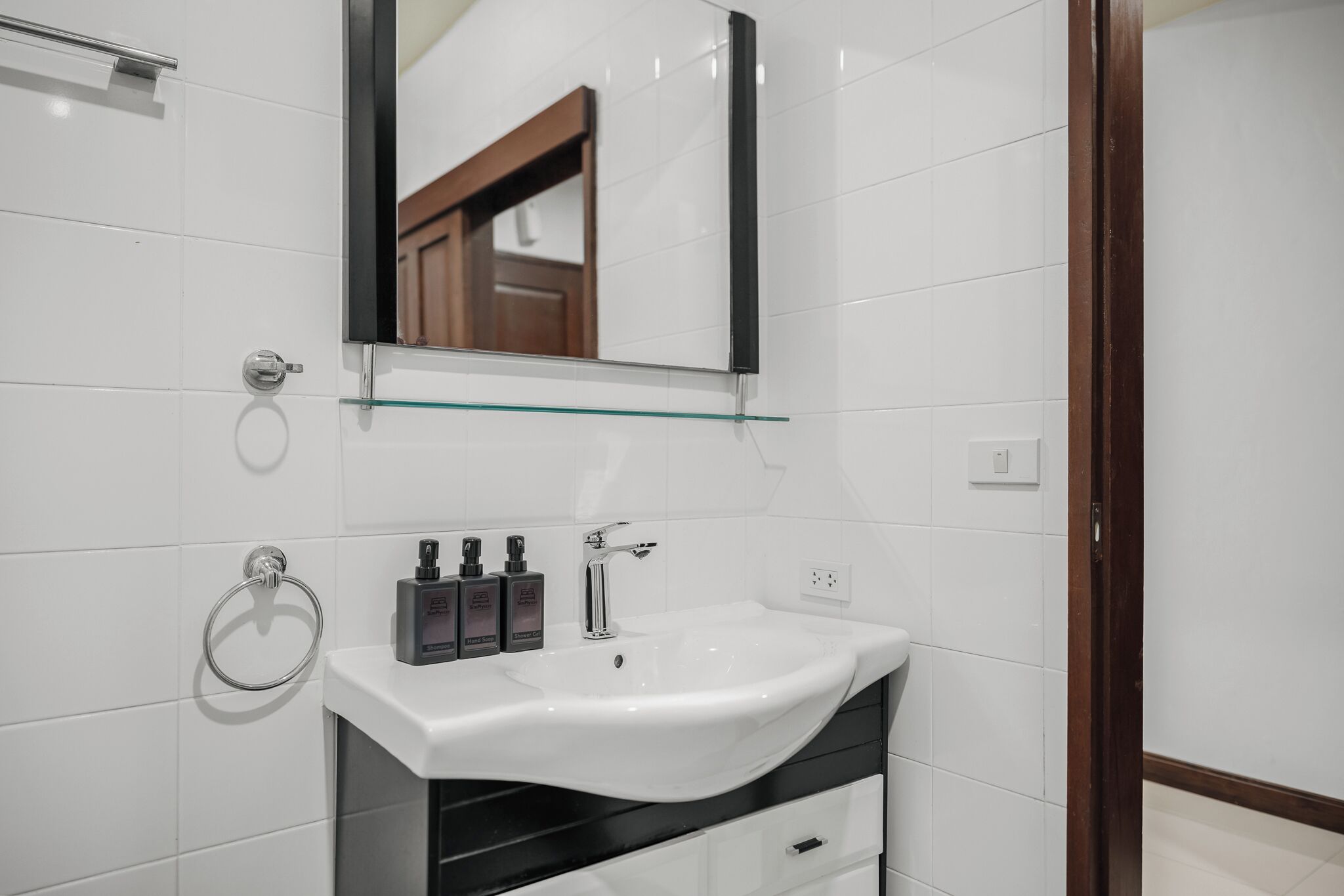 A modern bathroom with white tiles, a glass shelf above the sink, and a wooden door.