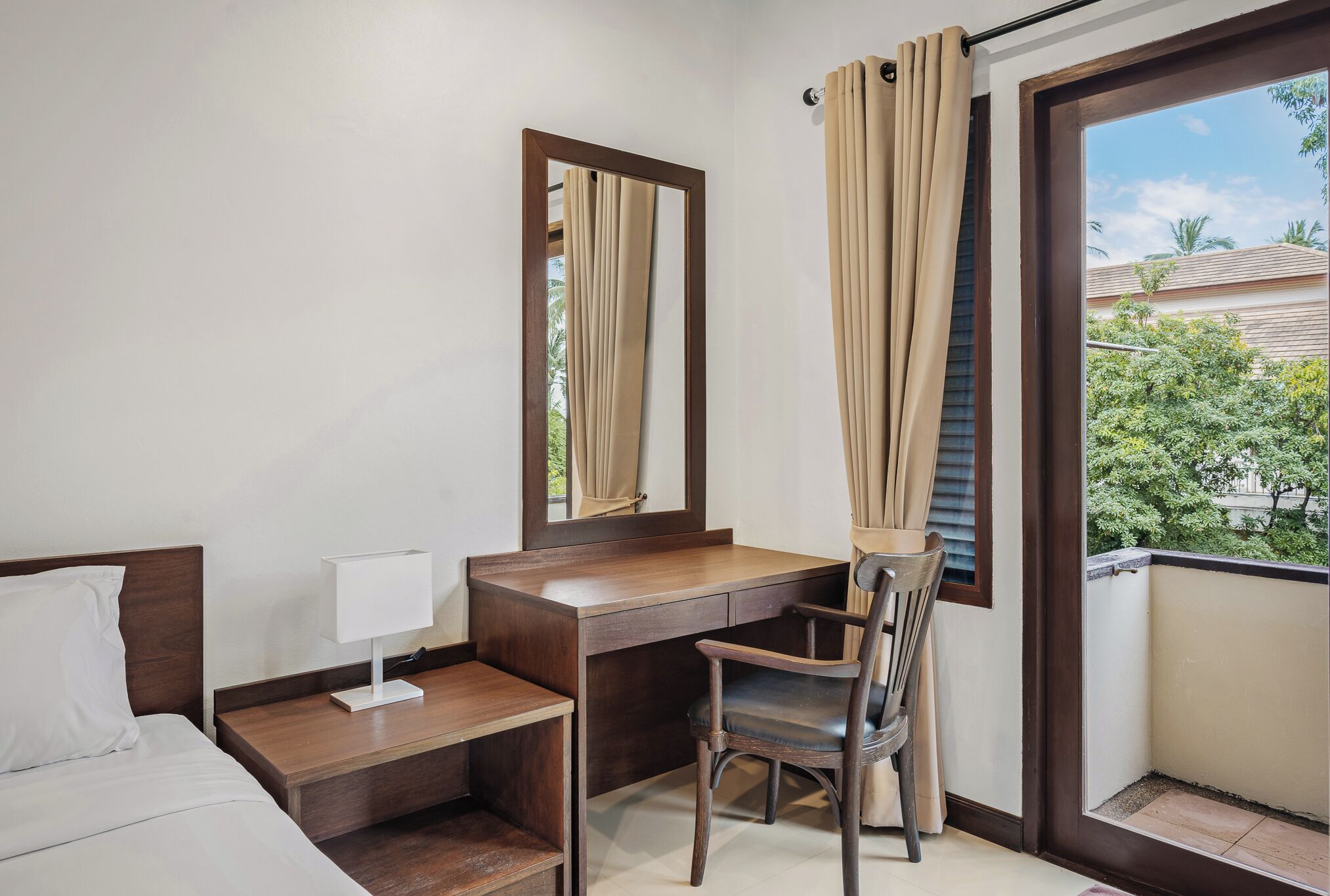 A compact hotel room with a bed, a desk, a chair, a mirror, and a window with curtains.