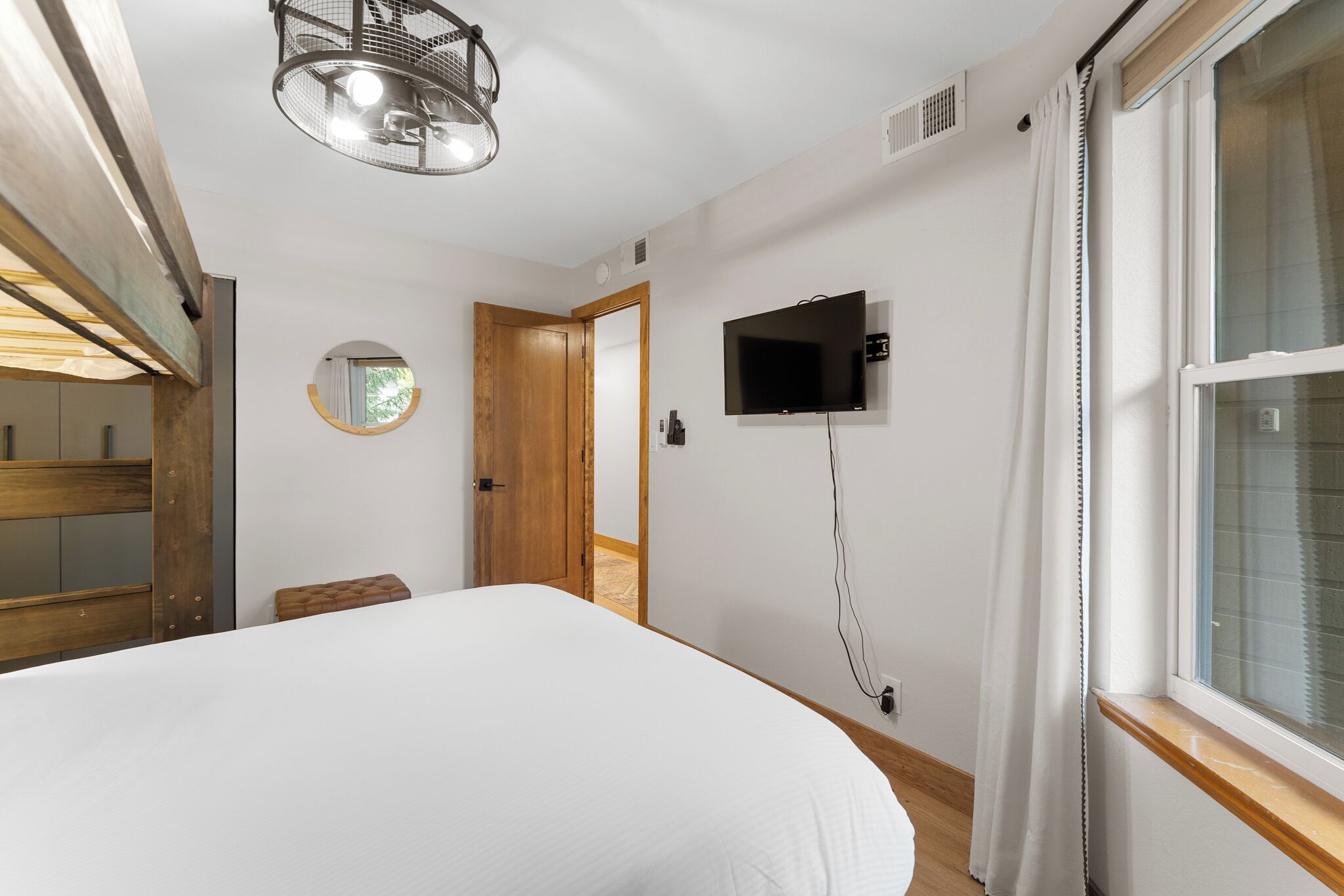 A bedroom with a bed, a TV mounted on the wall, a wooden door, and a window with curtains.