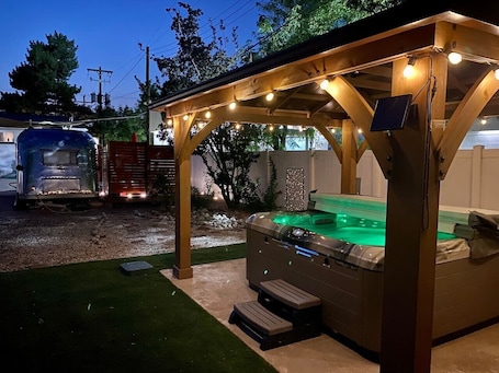 A hot tub under a wooden pergola with string lights, surrounded by a gravel area and a grassy lawn.. Year round:1959 Airstream Shiny Tiny Home HOTTuB