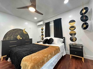 A bedroom with a bed, bedside tables, a ceiling fan, wall decorations, and an air conditioning unit.