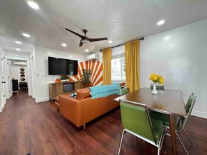A modern living room with a sofa, dining table, and chairs, a ceiling fan, and a TV mounted on the wall.