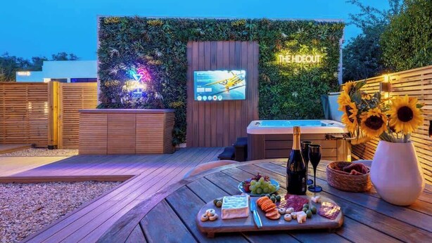 Un área de patio al aire libre con una mesa de madera dispuesta para una comida, un jacuzzi y una pantalla de televisión instalada en la pared.