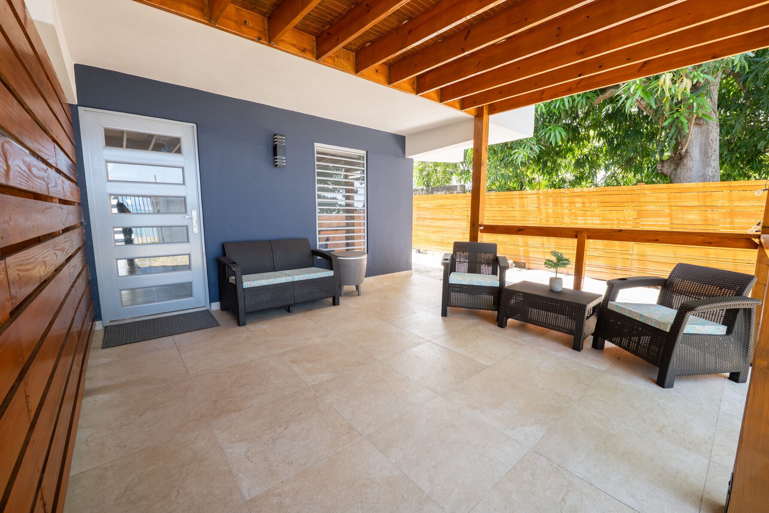 An outdoor patio with a wooden ceiling, a grey sofa, wicker chairs, and a small table.