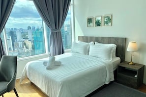Habitación de hotel con una cama grande, vistas a la ciudad, una silla gris y una mesita de noche con lámpara.