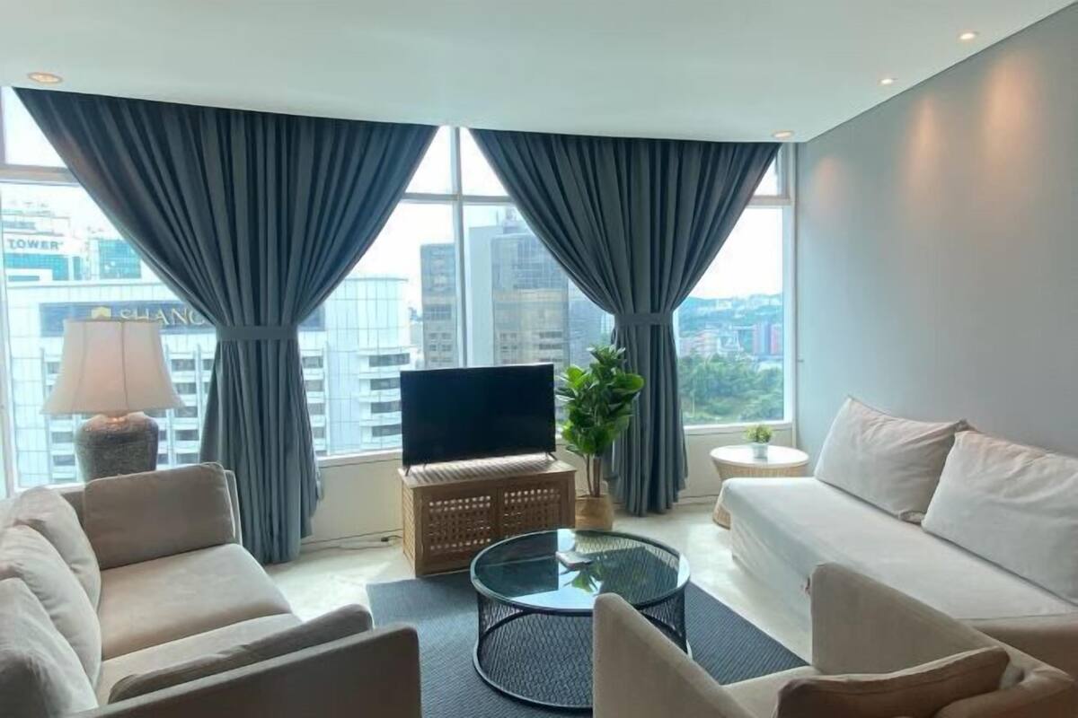A modern living room with a sofa, armchairs, a coffee table, a TV, and a view of the cityscape through large windows.