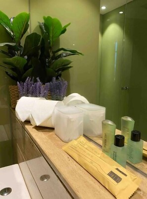 Un baño con pared verde, una planta en maceta, una toalla, un recipiente blanco y artículos de aseo.