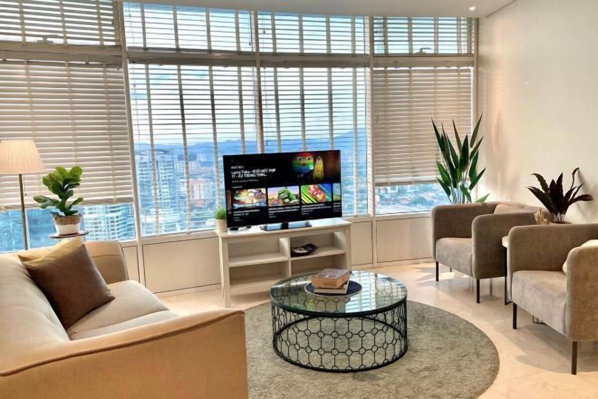 A modern living room with a sofa, armchairs, a glass coffee table, a TV on a stand, and large windows with blinds.