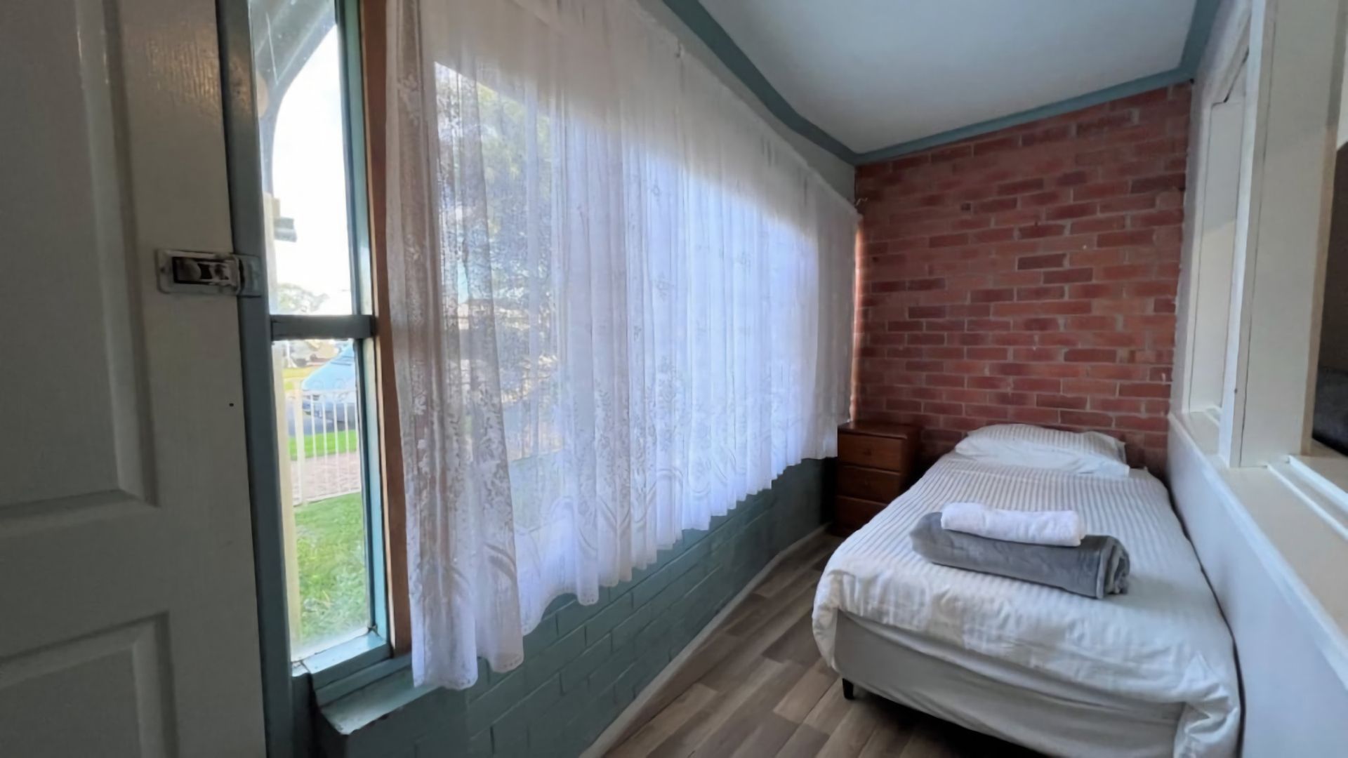 A room with a bed, a window with curtains, a brick wall, and a wooden floor.