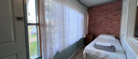A room with a bed, a window with curtains, a brick wall, and a wooden floor.