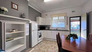 A compact kitchen with white cabinets, a refrigerator, an oven, and a dining table with chairs. - Cottage 1 ~ Come and relax by the river! (Sussex Inlet)