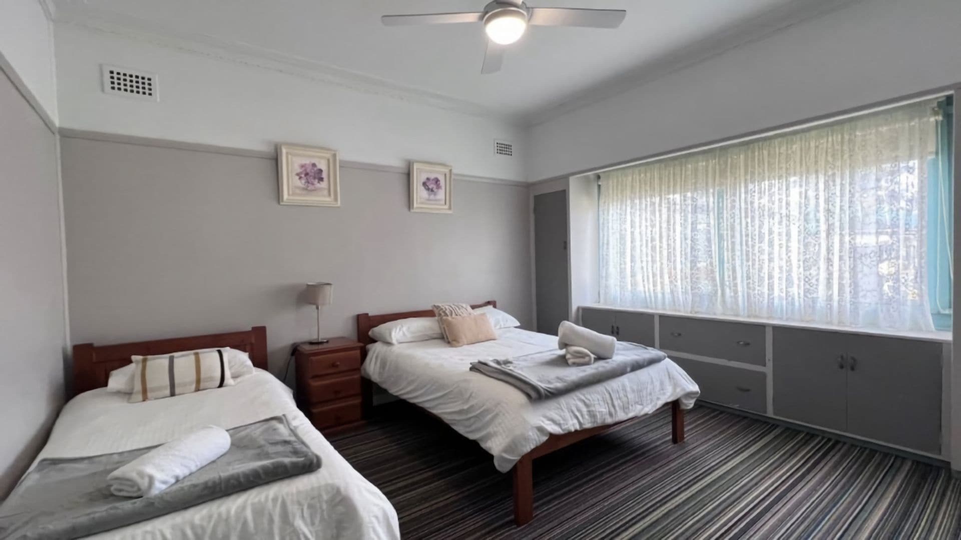 A hotel room with two single beds, a ceiling fan, a window with sheer curtains, and a small bedside table with a lamp.