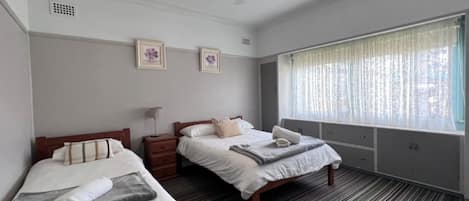 A hotel room with two single beds, a ceiling fan, a window with sheer curtains, and a small bedside table with a lamp.