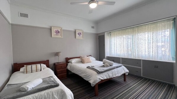 A hotel room with two single beds, a ceiling fan, a window with sheer curtains, and a small bedside table with a lamp.