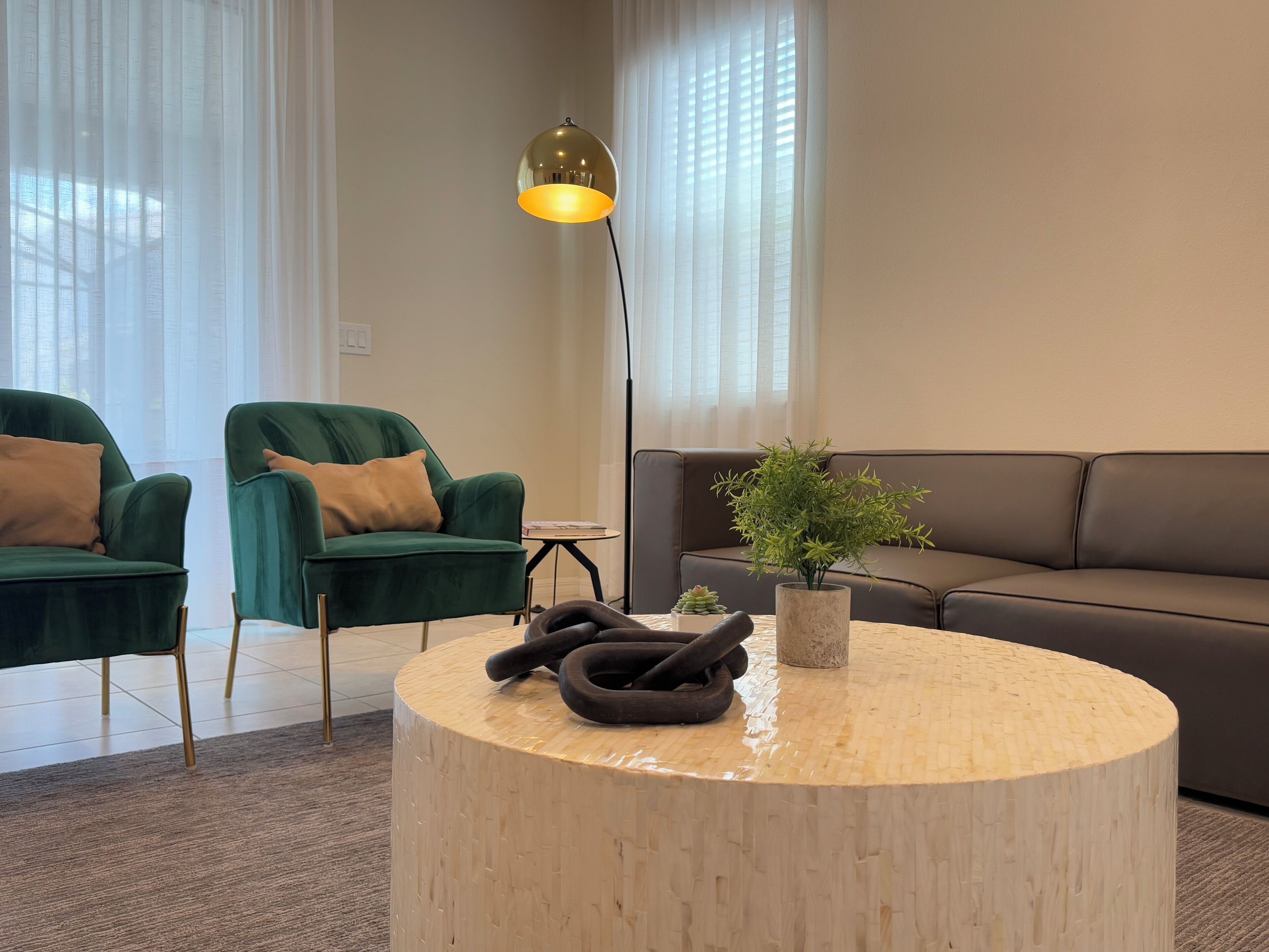 Een moderne woonkamer met twee groene fauteuils, een bruine bank, een ronde houten salontafel, een vloerlamp en een potplant.
