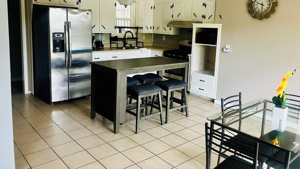 A modern kitchen with a stainless steel refrigerator, a central island with bar stools, white cabinetry, and a wall-mounted clock.