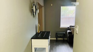 A narrow hallway with a white table and shelves, a window with blinds, and a dark-colored sofa in the background.