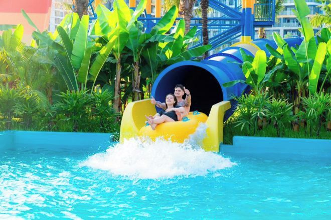 Un tobogán de agua con dos personas al final, rodeado de follaje y una piscina.