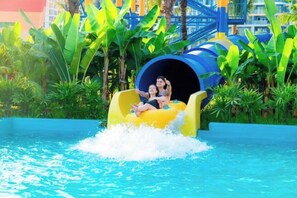 Eine Wasserrutsche mit zwei Personen am Ende, umgeben von Grünflächen und einem Schwimmbecken.